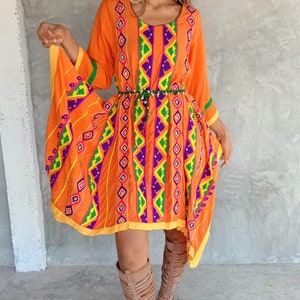 May include: A woman wearing a bright orange dress with colorful geometric embroidery and a beaded belt. The dress has a flowing, asymmetrical hemline. She is wearing brown leather gladiator sandals.