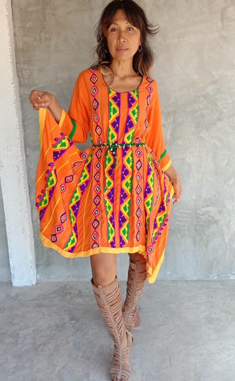 May include: A woman wearing a bright orange dress with colorful geometric embroidery and a beaded belt. The dress has a flowing, asymmetrical hemline. She is wearing brown leather gladiator sandals.