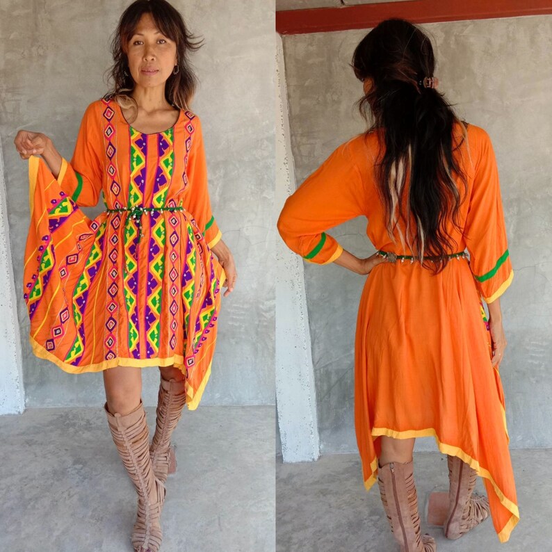 May include: A bright orange dress with a geometric pattern and a high-low hemline. The dress has a green and yellow trim and a purple and green embroidered design. The model is wearing the dress with a brown leather belt and brown leather boots.
