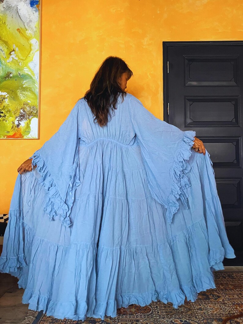 May include: A light blue maxi dress with long, flowing sleeves and tiered ruffles. The dress has a gathered waist and a full skirt. The model is standing in front of a yellow wall with a painting and a black door.