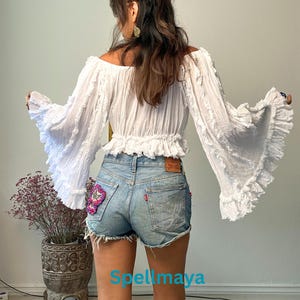 K&ouml;nnte beinhalten: Eine wei&szlig;e Off-Shoulder-Bluse mit langen, ger&uuml;schten &Auml;rmeln und einem verk&uuml;rzten Schnitt. Die Bluse wird mit hellblauen Jeansshorts kombiniert. Die Shorts haben einen dekorativen Aufn&auml;her auf der Ges&auml;&szlig;tasche. Der Text "Spellmaya" ist sichtbar.
