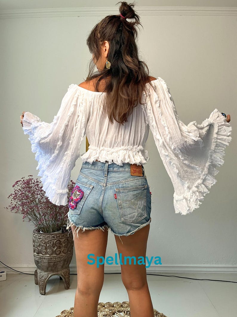 K&ouml;nnte beinhalten: Eine wei&szlig;e Off-Shoulder-Bluse mit langen, ger&uuml;schten &Auml;rmeln und einem verk&uuml;rzten Schnitt. Die Bluse wird mit hellblauen Jeansshorts kombiniert. Die Shorts haben einen dekorativen Aufn&auml;her auf der Ges&auml;&szlig;tasche. Der Text "Spellmaya" ist sichtbar.