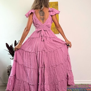 May include: A long, flowing, dusty rose dress with a tiered skirt and ruffled cap sleeves. The dress features a V-neckline and a tie at the waist. The model is holding the dress out, showcasing the full length.