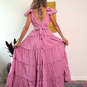 Pu&ograve; includere: Un lungo abito maxi a strati in un colore rosa polvere. L'abito ha una scollatura a V, maniche corte a sbuffo e una cintura da annodare in vita. L'abito &egrave; realizzato in un tessuto morbido e fluido.