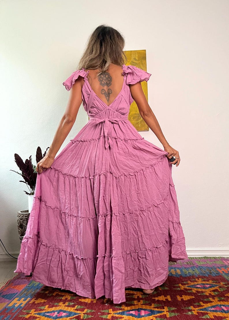 Pu&ograve; includere: Un lungo abito maxi a strati in un colore rosa polvere. L'abito ha una scollatura a V, maniche corte a sbuffo e una cintura da annodare in vita. L'abito &egrave; realizzato in un tessuto morbido e fluido.