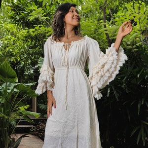 May include: A white cotton dress with a ruffled neckline and sleeves. The dress has a tie at the neckline and a gathered waist. The dress is knee-length and has a frayed hem.