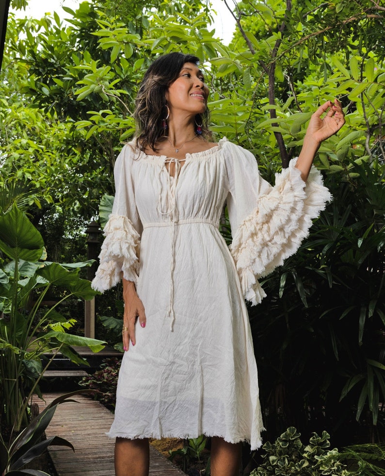 May include: A white cotton dress with a ruffled neckline and sleeves. The dress has a tie at the neckline and a gathered waist. The dress is knee-length and has a frayed hem.