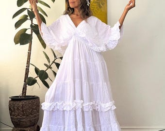 May include: A floor-length white dress featuring a V-neckline, an elasticized waist, and wide flutter sleeves. The skirt is constructed with multiple tiered layers, each finished with a ruffled hem. A white floral headpiece is worn on the head.