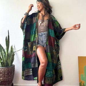 May include: A long, open-front kimono featuring a patchwork design of various green, black, and maroon fabric panels with intricate patterns. The garment has wide sleeves and two front pockets. The text JK3 is visible in the top left corner.