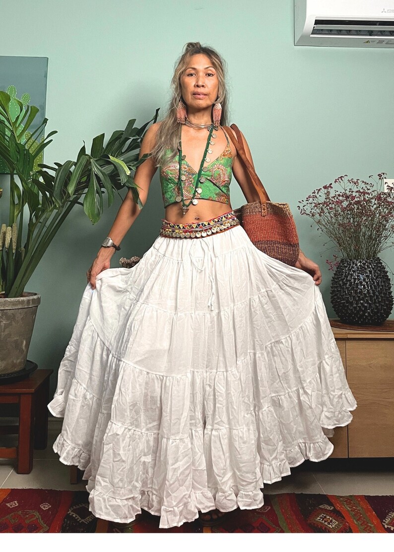 K&ouml;nnte beinhalten: Eine Frau tr&auml;gt ein gr&uuml;nes und goldenes gemustertes Crop-Top, einen wei&szlig;en, geschichteten Rock und eine braune gewebte Tasche. Sie tr&auml;gt einen silbernen G&uuml;rtel mit bunten Perlen und Charms.
