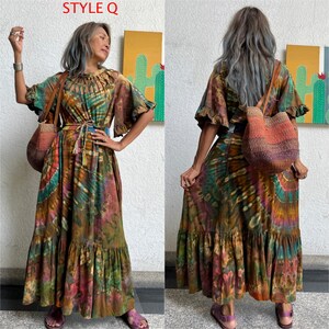 May include: A woman wearing a long, flowing, multi-colored tie-dye dress with a brown leather belt and a brown woven tote bag. The dress has a ruffled neckline and a tiered skirt. The woman is standing in front of a wall with a yellow and green painting.