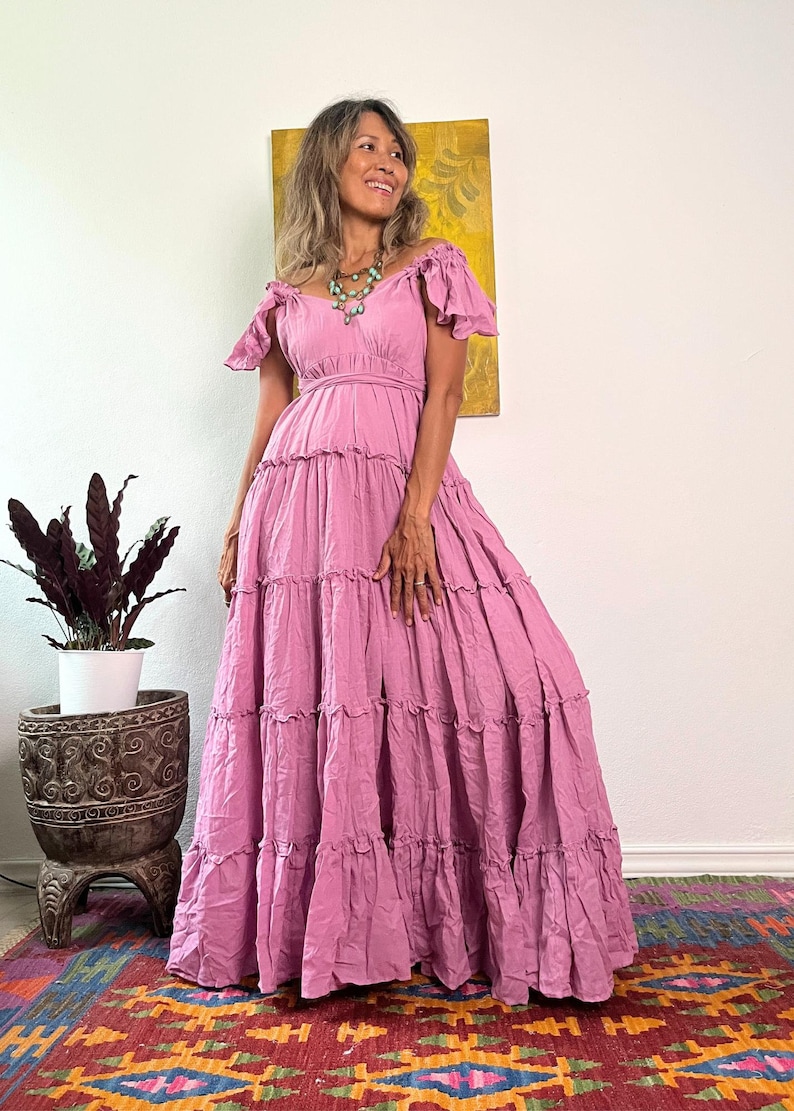 May include: A long, flowing, dusty rose-colored dress with off-the-shoulder sleeves and a tiered skirt. The dress features a fitted bodice and a full, ruffled skirt. A turquoise necklace adds a pop of color. The dress is suitable for a special occasion.