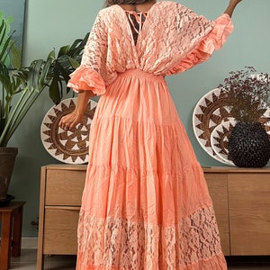 Puede incluir: Un vestido maxi de color melocot&oacute;n con una superposici&oacute;n de encaje y una falda de capas. El vestido tiene un escote en V y un lazo en la espalda.