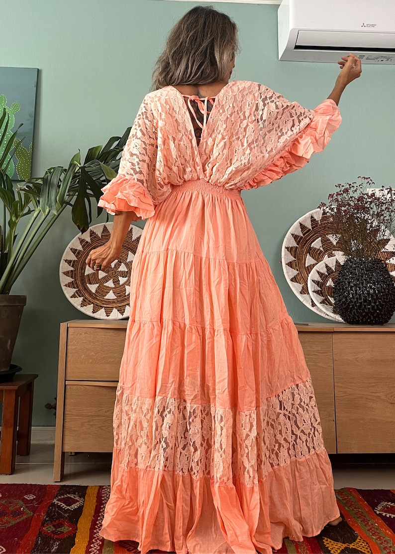 Puede incluir: Un vestido maxi de color melocot&oacute;n con una superposici&oacute;n de encaje y una falda de capas. El vestido tiene un escote en V y un lazo en la espalda.