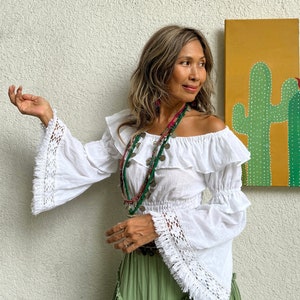 May include: A white off-the-shoulder blouse with crochet trim and bell sleeves. The blouse has a ruffled neckline and a fitted bodice. The model is wearing a green skirt and multiple necklaces.