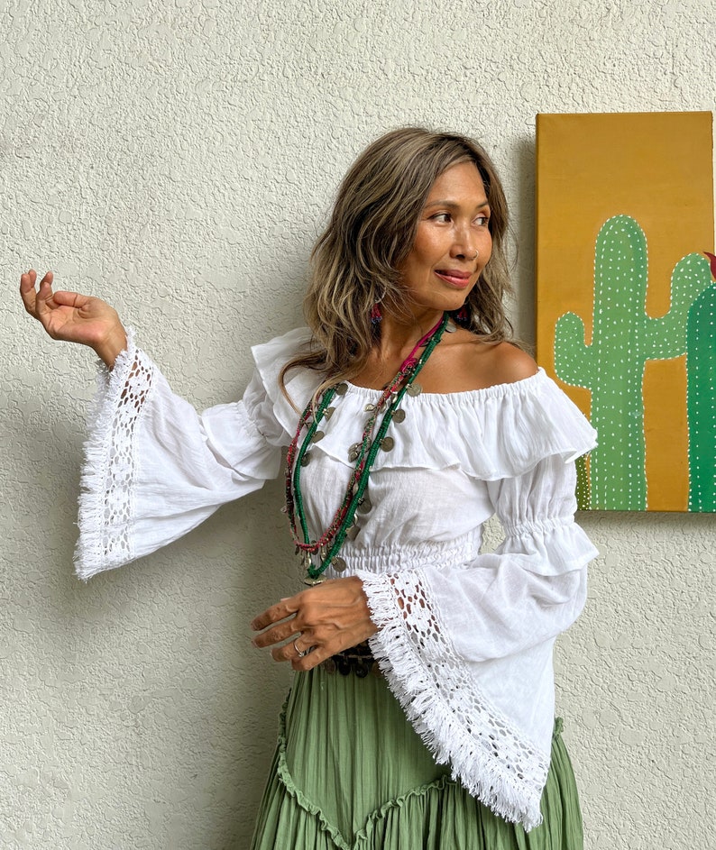 May include: A white off-the-shoulder blouse with crochet trim and bell sleeves. The blouse has a ruffled neckline and a fitted bodice. The model is wearing a green skirt and multiple necklaces.