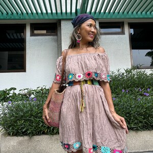 May include: A woman wearing a brown off-the-shoulder dress with crocheted trim, a colorful belt, and a blue bandana. She is holding a woven bag and standing in front of a building with a green pergola.