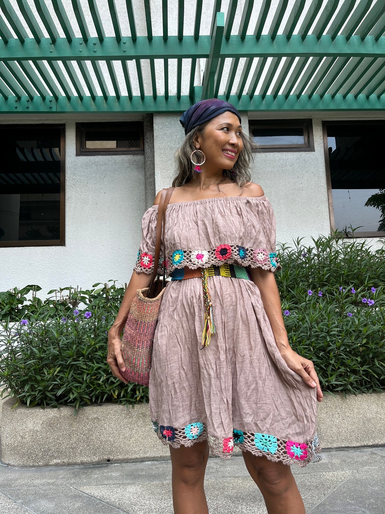 May include: A woman wearing a brown off-the-shoulder dress with crocheted trim, a colorful belt, and a blue bandana. She is holding a woven bag and standing in front of a building with a green pergola.