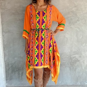 May include: A woman wearing a bright orange dress with colorful geometric embroidery and a green beaded belt. She is wearing brown leather knee-high boots.