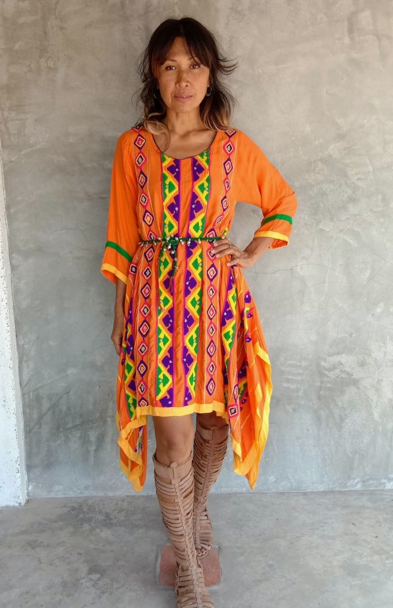 May include: A woman wearing a bright orange dress with colorful geometric embroidery and a green beaded belt. She is wearing brown leather knee-high boots.