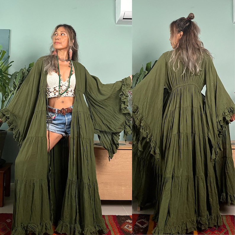 Op de afbeelding: Olijfgroene maxi-jurk met gelaagde ruches en wijde mouwen. De jurk heeft een vloeiende, bohemien stijl, ideaal om te dragen in laagjes. De jurk is vloerlang en heeft een striksluiting in de taille. De mouwen hebben een gefranjerde rand.