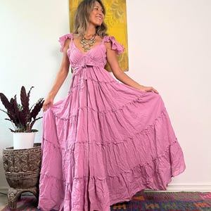 May include: A flowing, dusty rose maxi dress with a tiered skirt and ruffled cap sleeves. The dress features a V-neckline and a tie at the waist. The model is wearing a turquoise necklace. The background includes a colorful rug and a potted plant.