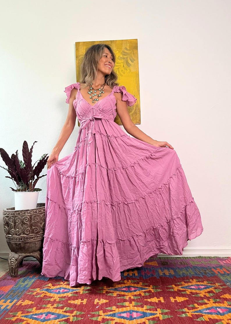 May include: A flowing, dusty rose maxi dress with a tiered skirt and ruffled cap sleeves. The dress features a V-neckline and a tie at the waist. The model is wearing a turquoise necklace. The background includes a colorful rug and a potted plant.
