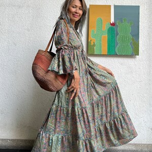 May include: A woman wearing a long, tiered floral dress with bell sleeves and a brown woven tote bag. She is standing in front of a wall with a painting of cacti on it.