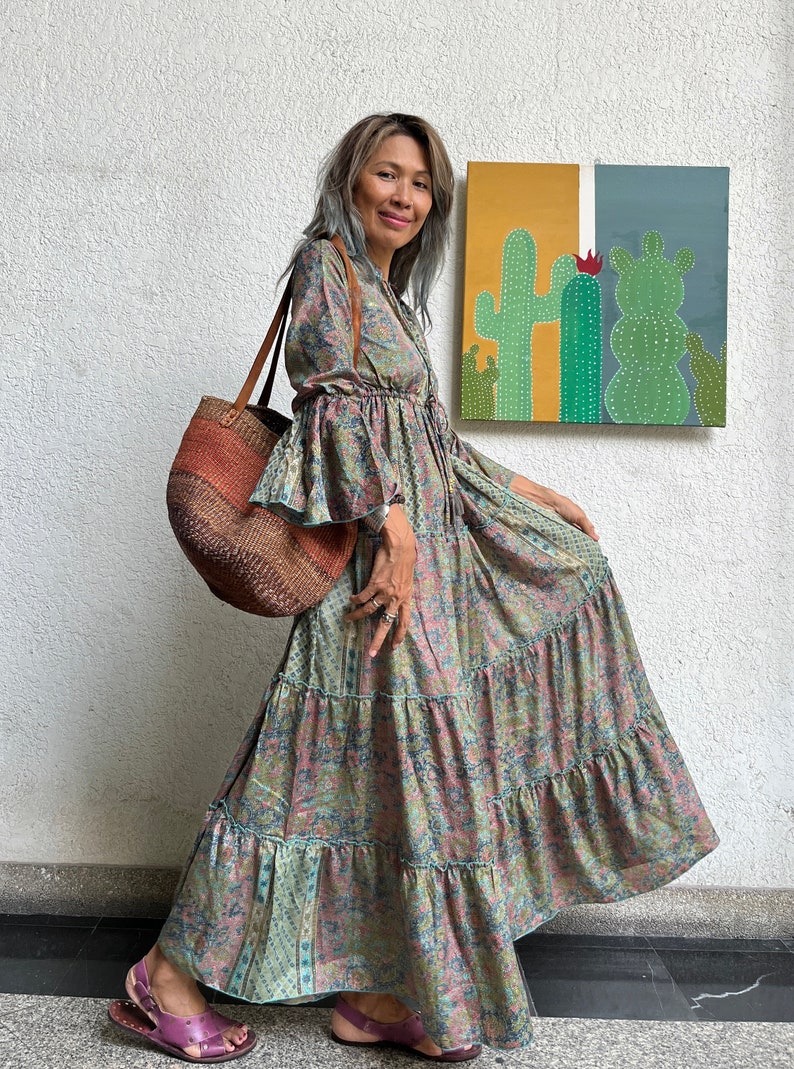 May include: A woman wearing a long, tiered floral dress with bell sleeves and a brown woven tote bag. She is standing in front of a wall with a painting of cacti on it.