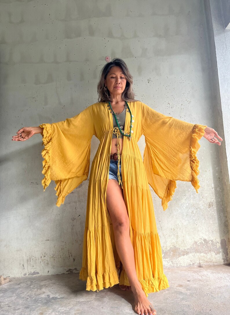May include: A flowing, mustard yellow maxi dress with tiered skirt and wide, ruffled sleeves. The dress is open in the front and tied at the waist. A green necklace is visible. The dress is made of a lightweight fabric.