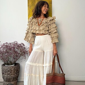 Pode incluir: Uma mulher usando um top cropped bege com babados e cal&ccedil;as brancas de perna larga. Ela est&aacute; segurando uma bolsa tote tecida marrom e vermelha com al&ccedil;as de couro. Um colar e um vaso decorativo com uma planta tamb&eacute;m s&atilde;o vis&iacute;veis.