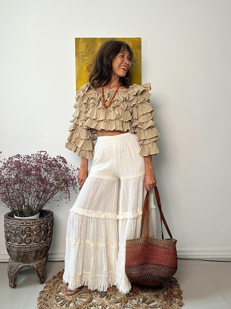 Pode incluir: Uma mulher usando um top cropped bege com babados e cal&ccedil;as brancas de perna larga. Ela est&aacute; segurando uma bolsa tote tecida marrom e vermelha com al&ccedil;as de couro. Um colar e um vaso decorativo com uma planta tamb&eacute;m s&atilde;o vis&iacute;veis.