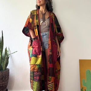 May include: A long, open-front kimono robe featuring a patchwork design with various fabric squares in shades of red, orange, yellow, green, and brown. The garment includes two large front pockets and wide sleeves, worn over a neutral top and denim shorts.