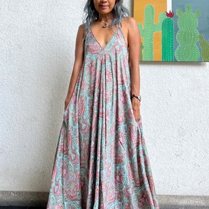 May include: A woman wearing a long, flowing maxi dress with a paisley print in shades of teal, pink, and brown. The dress has a V-neckline and thin straps. She is standing in front of a white wall with a painting of cacti on it.