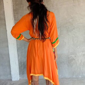May include: A long-sleeved, orange dress with yellow and green trim. The dress has a high neckline and a flowing, asymmetrical hemline. The dress is cinched at the waist with a brown belt.