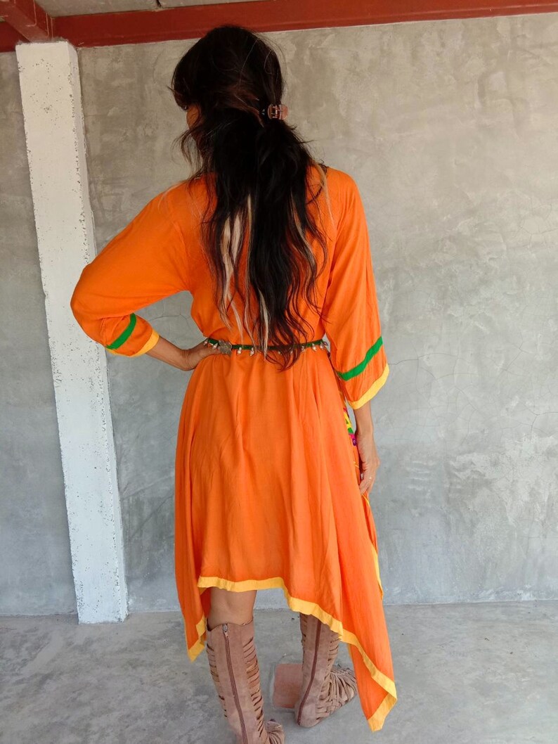 May include: A long-sleeved, orange dress with yellow and green trim. The dress has a high neckline and a flowing, asymmetrical hemline. The dress is cinched at the waist with a brown belt.