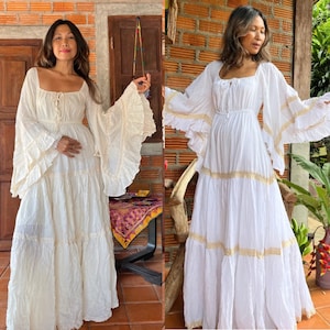 May include: Two women wearing white maxi dresses with lace trim and long sleeves. The dresses have a tiered design and are made of a lightweight fabric.