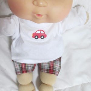 May include: A doll with light skin, brown eyes, and light hair wearing a white long-sleeve shirt featuring a small red car graphic on the chest and plaid shorts with red, white, and black patterns. The doll has a round face and visible toes.