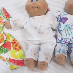 May include: Two dolls wearing pajamas. One doll is wearing white pajamas with a repeating pattern of a bear holding a present. The other doll is wearing white pajamas with a blue and purple sea life pattern. A pair of fruit-patterned pajamas is on the left.