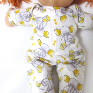 May include: A Cabbage Patch Kid doll wearing a white and yellow pajamas with a gray elephant print. The pajamas have yellow accents on the elephants.