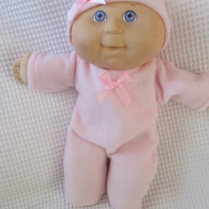 May include: A pink baby doll wearing a pink fleece sleeper and a pink hat with a pink bow. The doll has blue eyes and a rosy complexion.