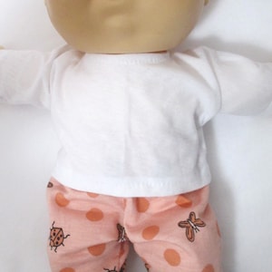 May include: A Cabbage Patch Kid doll wearing a white t-shirt and pink and brown ladybug and butterfly print pants.