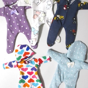 May include: A collection of doll clothes, including several hooded onesies and matching hats. The onesies feature various patterns, such as polka dots, hearts, and teddy bears. The color palette includes purple, white, blue, and rainbow hues.