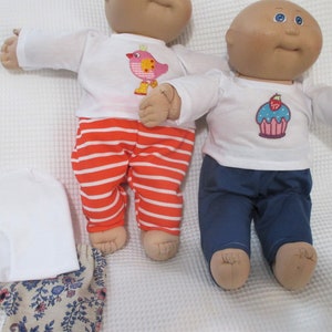 May include: Two Cabbage Patch Kids dolls wearing different outfits. One doll is wearing a white shirt with a pink bird design and red and white striped pants. The other doll is wearing a white shirt with a cupcake design and blue pants.