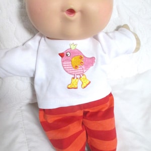 May include: A doll with a round face, brown eyes, and light skin wearing a white long-sleeve shirt featuring an embroidered pink bird with a yellow crown and yellow boots, paired with orange and red horizontally striped pants.