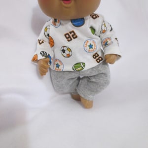 May include: A Cabbage Patch Kid doll wearing a white long-sleeved shirt with sports ball graphics and the numbers 92 and 88. The doll is also wearing gray sweatpants. The doll has brown skin, rosy cheeks, and brown eyes.