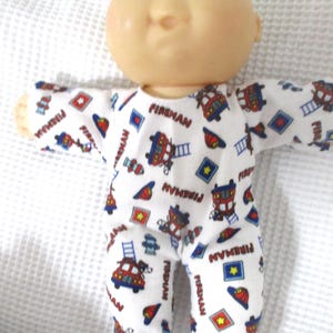 May include: A Cabbage Patch Kid doll wearing a white and red fireman-themed sleeper with the word "Fireman" repeated throughout the design.