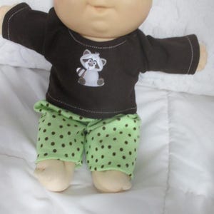 May include: A doll with a round head, light skin, and blue eyes wearing a dark brown long-sleeved shirt featuring a small graphic of a raccoon on the chest. The doll is also wearing light green pants with a pattern of small dark brown polka dots.