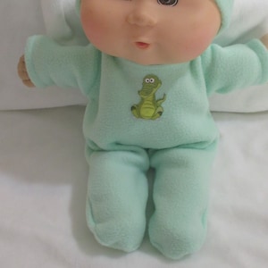 May include: A green plush baby doll wearing a green hat and a green onesie with a green alligator graphic. The doll has a round face with brown eyes and rosy cheeks.