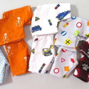 May include: Six pairs of pajamas for dolls, featuring various patterns and colors. The pajamas include a blue and white plaid, orange with white skeletons, white with traffic signs and a construction vehicle, white with traffic signs, white with red and orange geometric shapes, and red with white paw prints and hearts.